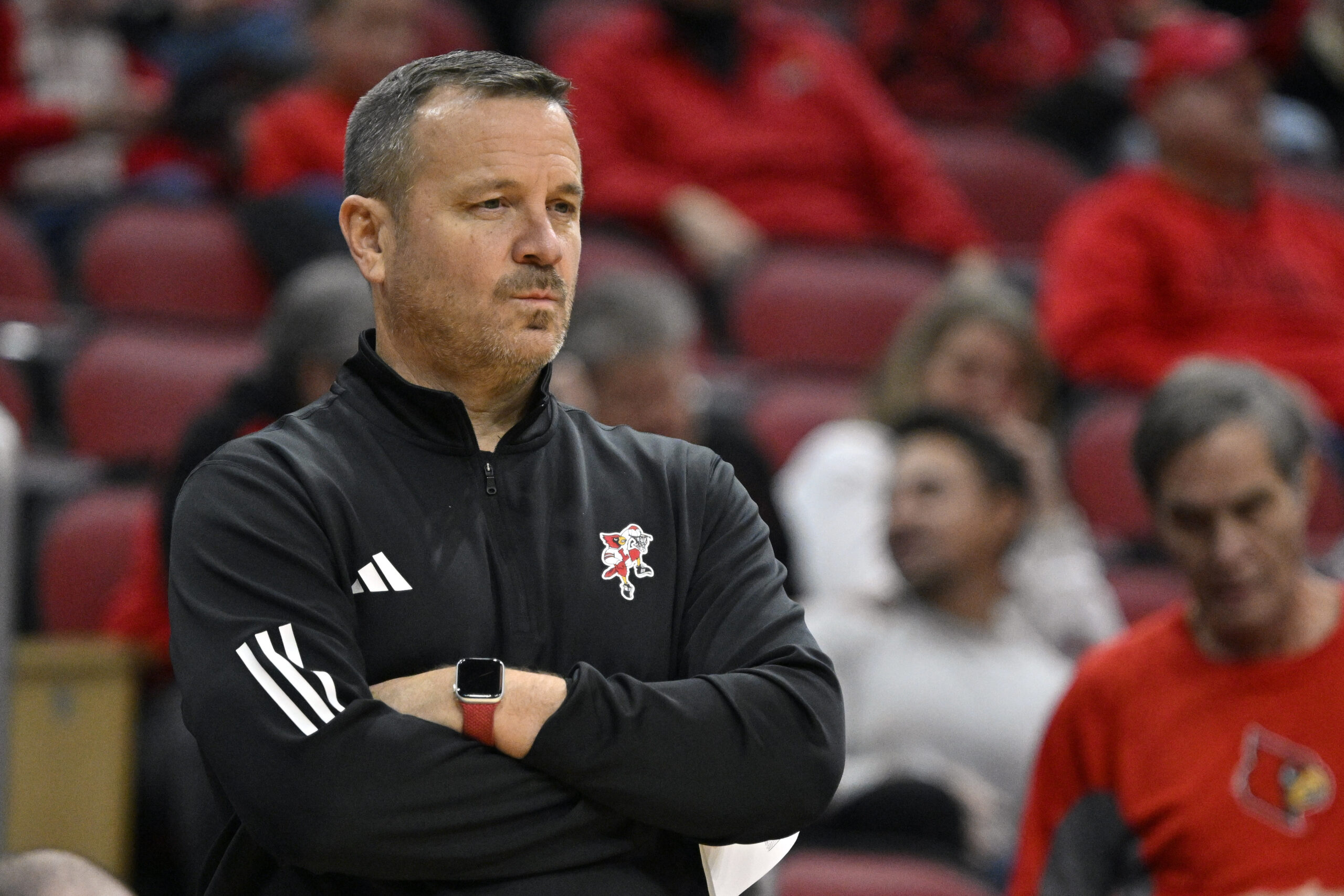 Louisville head coach Jeff Walz appears in focus in the foreground, wearing a black Adidas quarterzip with his arms crossed.