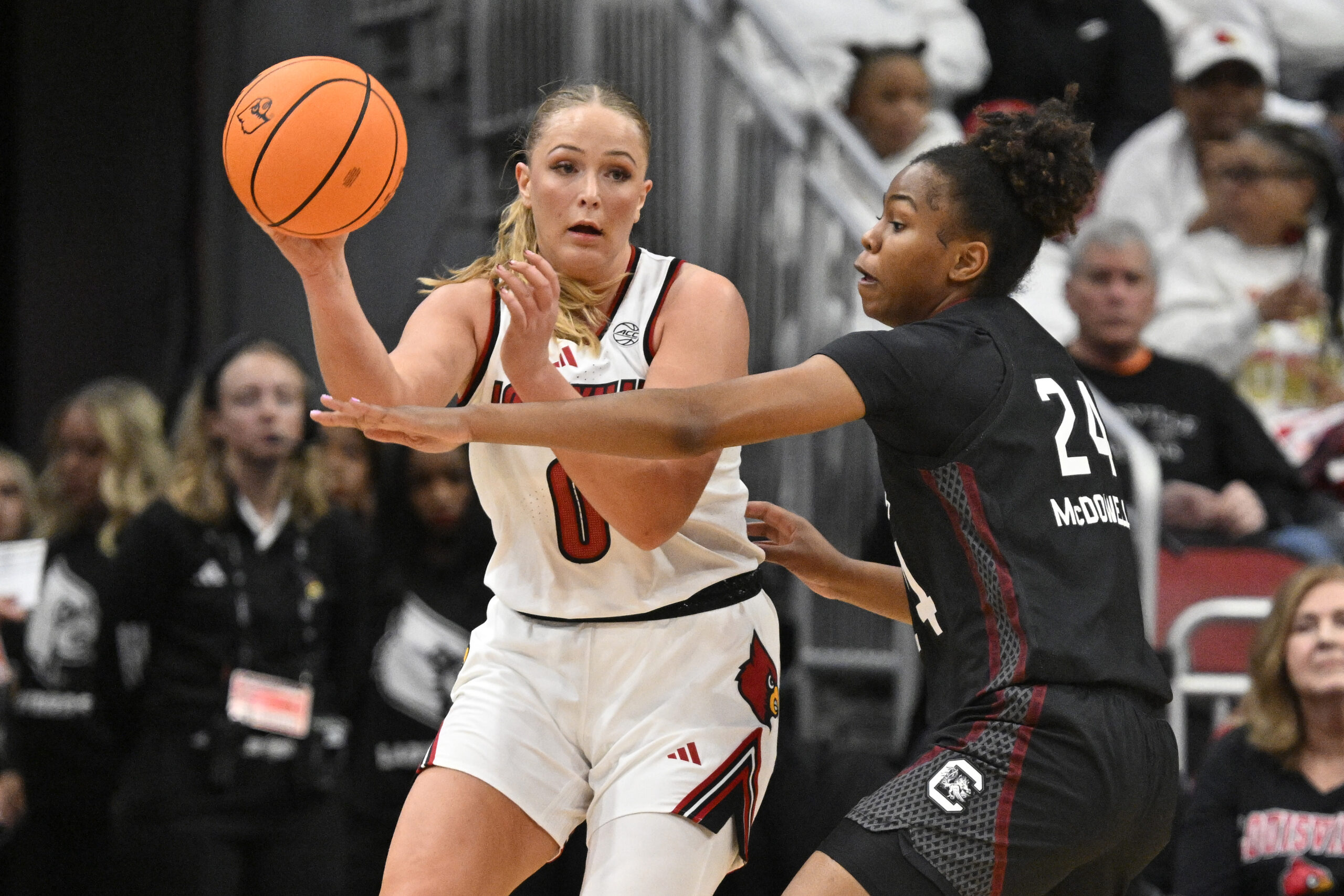 Laura Ziegler (0) wearing a white Louisville Cardinals jersey, is captured mid-motion attempted to pass the ball around the outstretched hand of South Carolina's Ayla McDowell (24), wearing a black jersey.