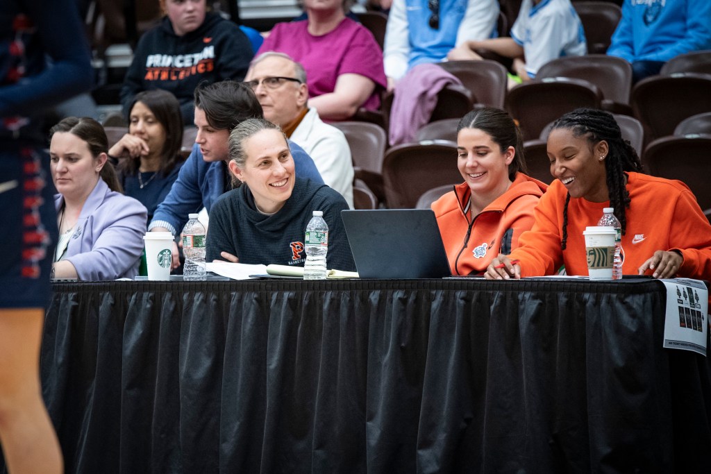 Princeton head coach Carla Berube and assistant coaches Lauren Dillon and Jordan Edwards sit courtside to scout an opponent. They are all smiling, and Dillon and Edwards are looking down at a laptop.