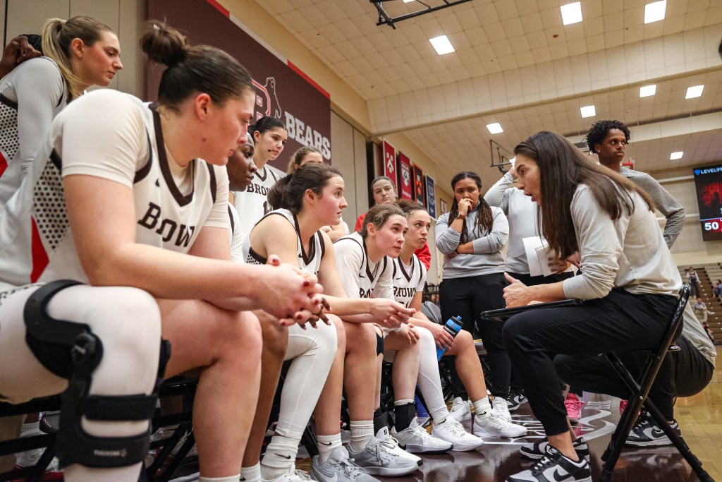 Brown head coach Monique LeBlanc sits in a chair in front of her bench and talks to her team during a huddle. She is gesturing with her left hand, and her players are looking at her as she speaks.