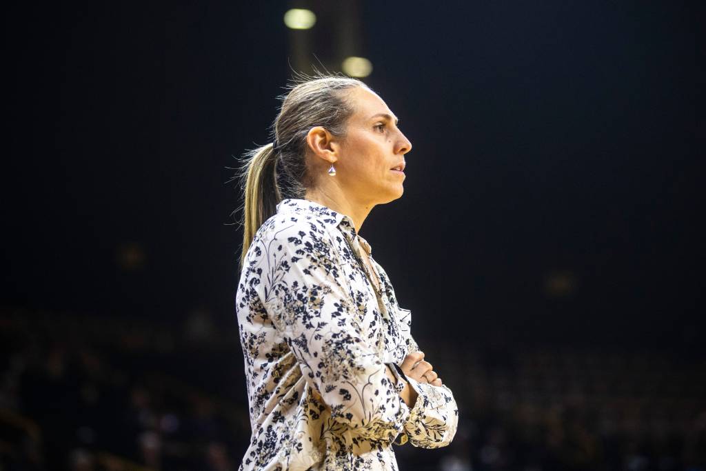 Princeton head coach Carla Berube is shown from the side and from the waist up. She has her arms crossed over her chest and a neutral expression on her face. The background behind her is black, contrasting with her light floral shirt.