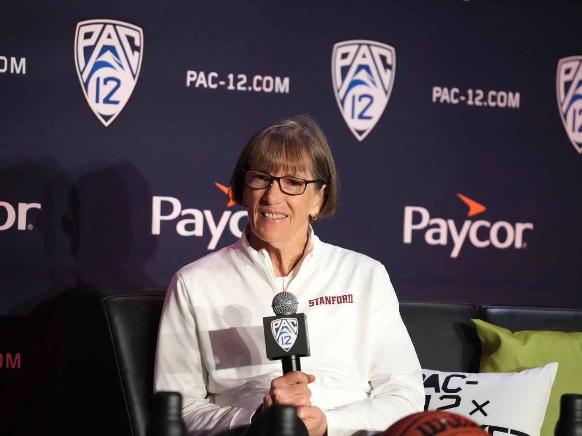 Stanford Cardinal head coach Tara VanDerveer sits and holds a microphone as she speaks at a preseason media day. Pac-12 logos are visible on the backdrop behind her.