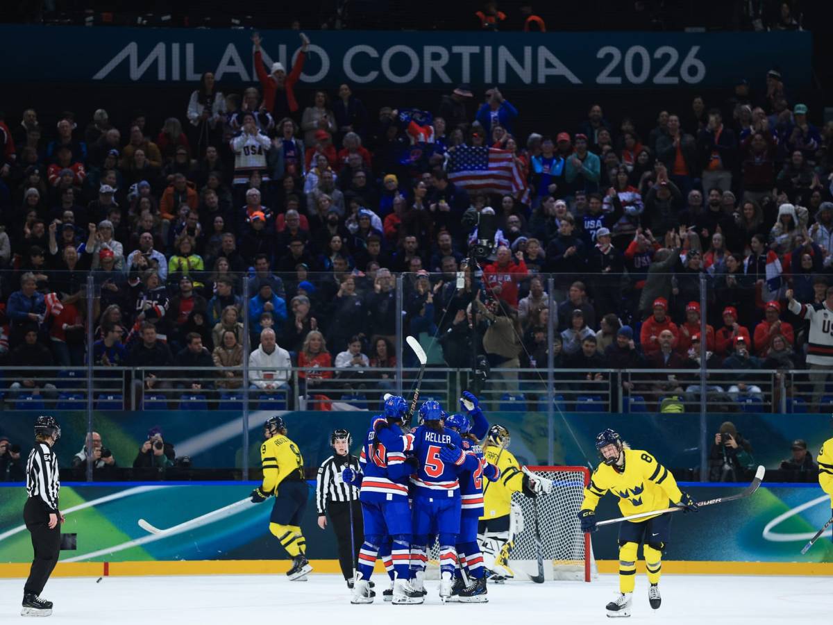 Team USA celebrates their goal with a group hug in front of the net. They are wearing blue home uniforms, while dejected looking Swedish players dot the background in yellow away ones.