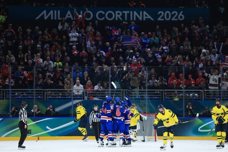 Team USA celebrates their goal with a group hug in front of the net. They are wearing blue home uniforms, while dejected looking Swedish players dot the background in yellow away ones.