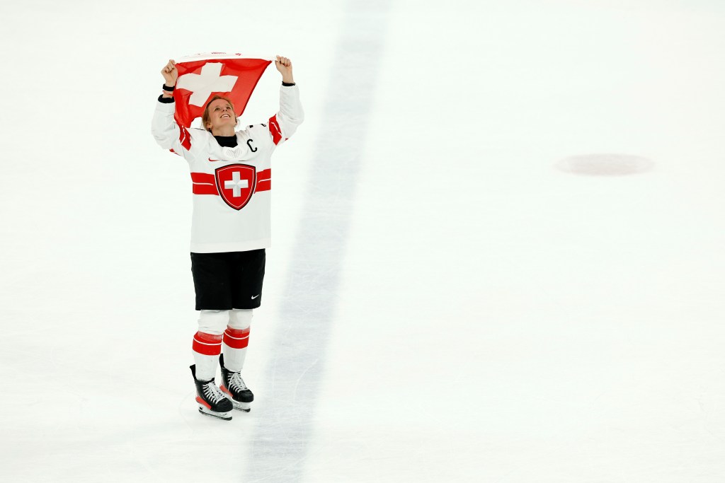 Captain Lara Christen holds a Swiss flag above her head while skating over the blue line and looking up to the sky