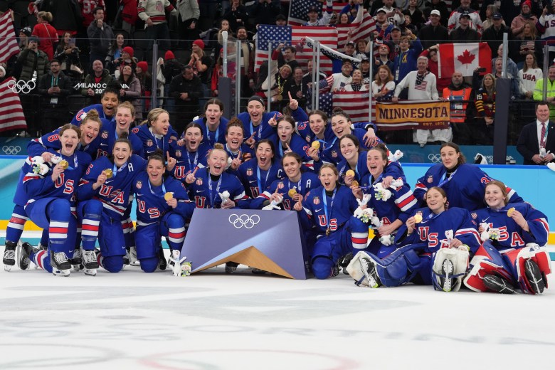 Team USA piles together and smiles with their gold medal after winning the 2026 Olympics. They are all wearing blue home uniforms.