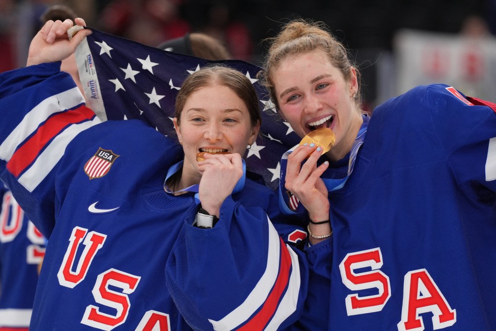Frankel and Harvey smile and bite their medals while holding a U.S. flag behind their backs. They are wearing blue home uniforms.