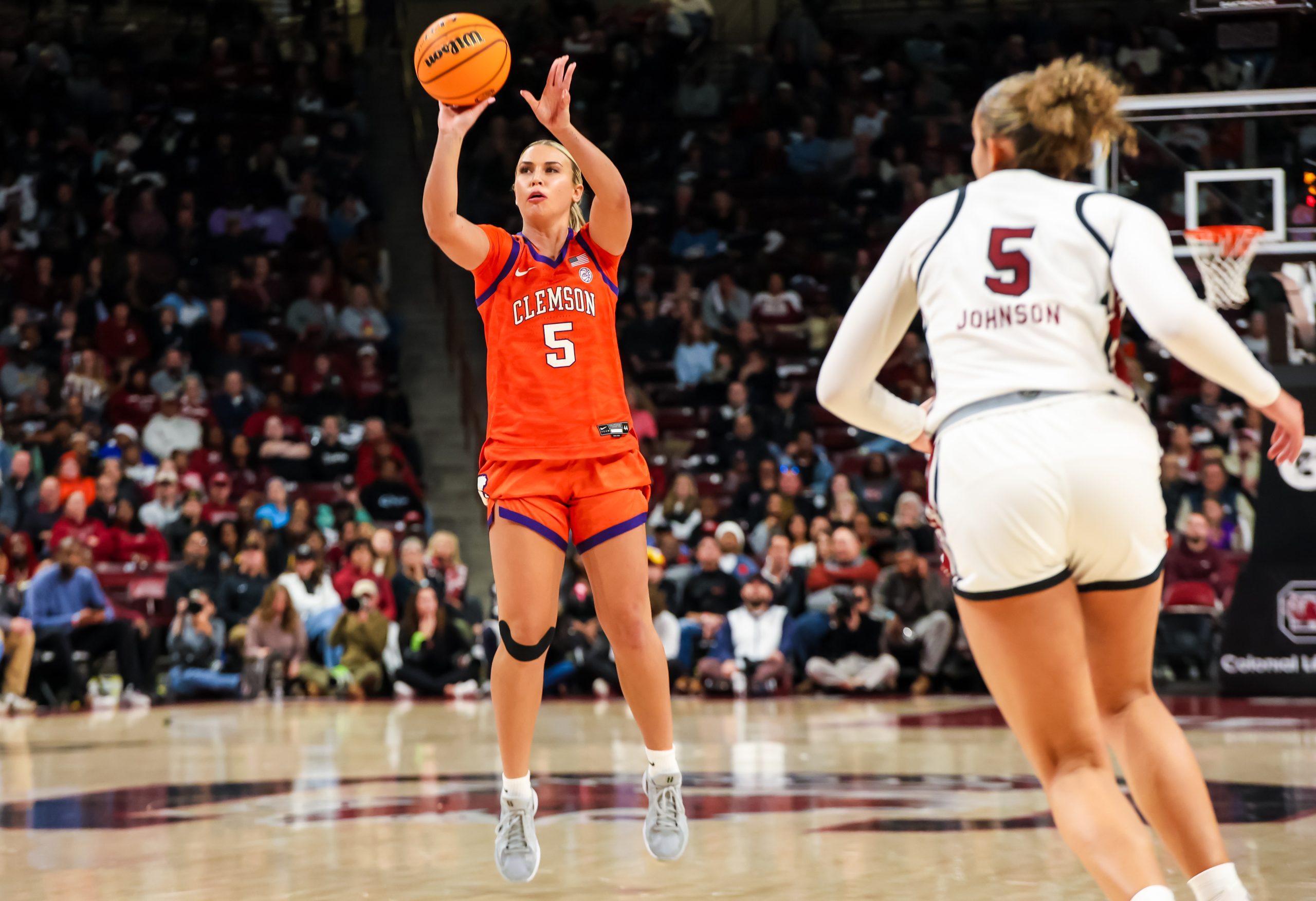 Clemson guard Hannah Kohn, wearing a bright orange jersey, is captured just as she releases a 3-point shot as South Carolina guard Tessa Johnson, wearing a white Gamecock jersey, moves in the foreground. 