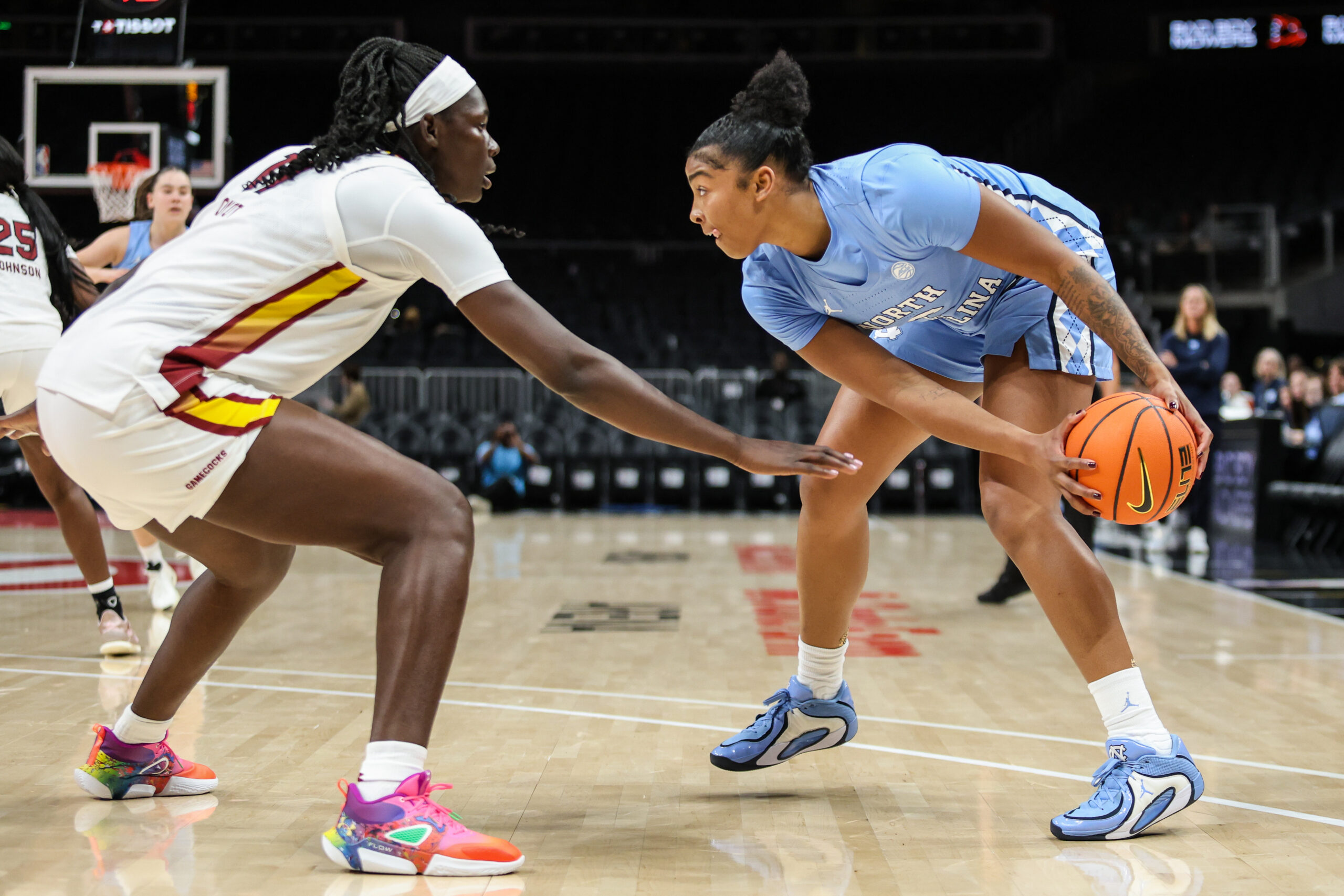 North Carolina forward Nyla Harris, wearing a baby blue Tar Heels jersey, holds the ball low and away from South Carolina's Madina Okot, wearing a white Gamecocks jersey. Okot has her arms out wide to keep Harris in place.