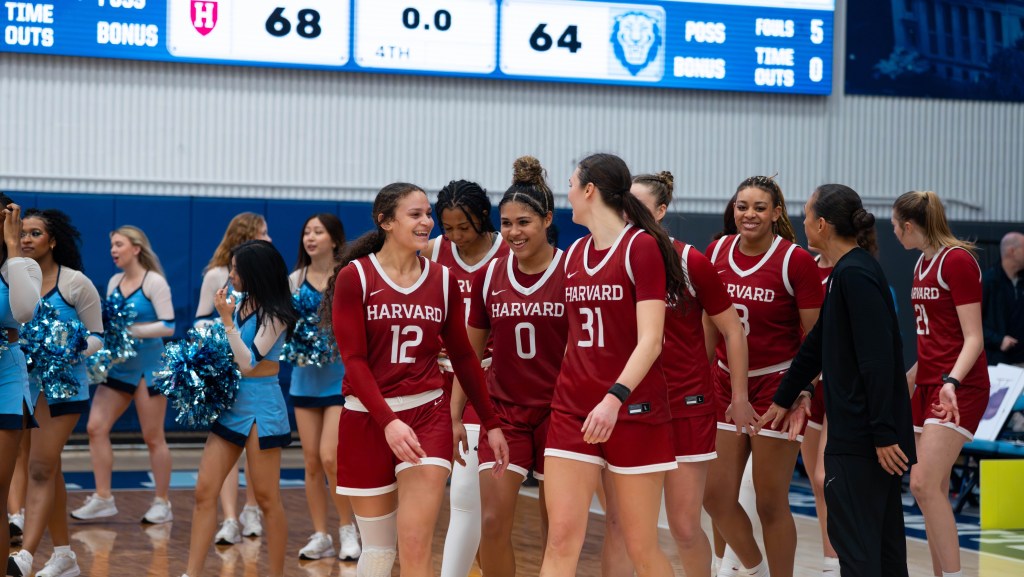 Harvard players and head coach Carrie Moore walk off the court together after a win over Columbia. Many of them are smiling and talking to each other. The bottom of the scoreboard, showing a 68-64 score and 0.0 seconds left, is visible in the background.