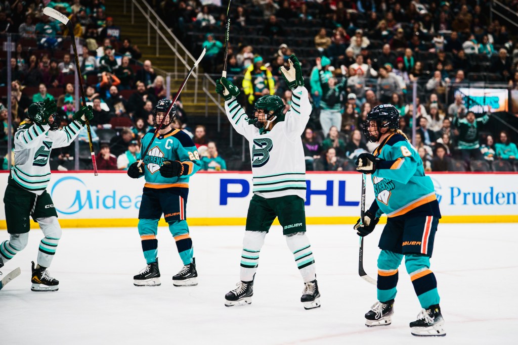 Huber (in white) smiles and raises her arms in celebrate. One of her teammates is skating over to her, as two Sirens players (in teal) stand dejected on either side of her.