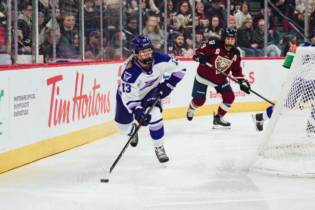 Zumwinkle skates behind the net with the puck, looking up ice to assess her options. She is wearing a white away uniform.
