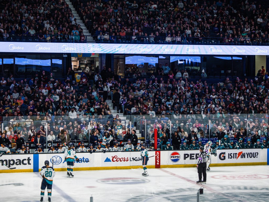 Some Sirens stand on the ice during a TV timeout, while others sit on the bench. The Torrent sit on the bench to their left. The Sirens are in white away uniforms, while the Torrent are in teal ones.