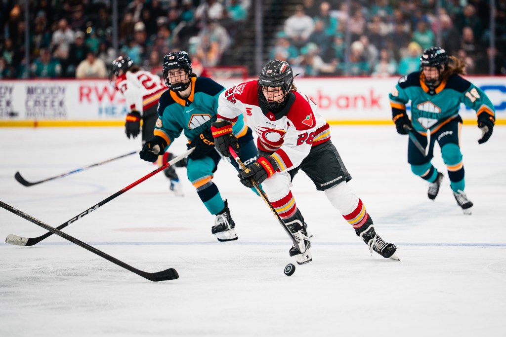 Clark (in white) looks down as she skates with the puck, with New York players (in teal) closing in around her.