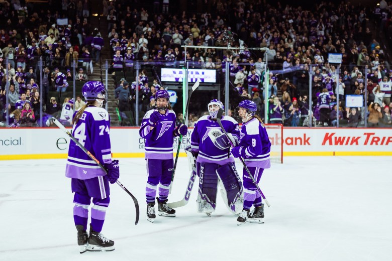 Four Frost players smile as they celebrate their win with a roaring crowd in the background. They are wearing purple home uniforms.