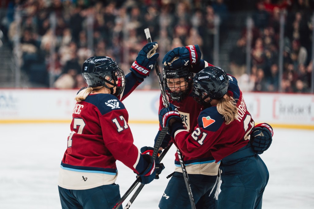 Three Victoire players smile and celebrate a goal with a group hug. They are wearing maroon home uniforms.