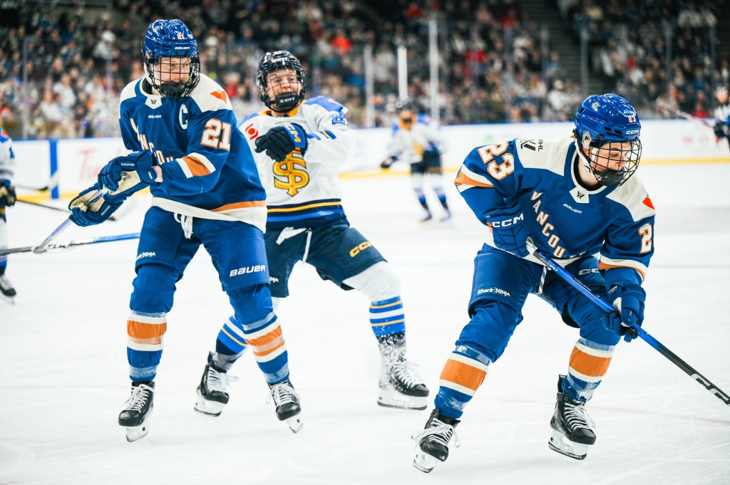One Goldeneyes player appears to have secured the puck, which is out of frame, as a Vancouver and Toronto player continue to battle in the background. The Goldeneyes are wearing blue home uniforms, while the Sceptres is in a white away one.
