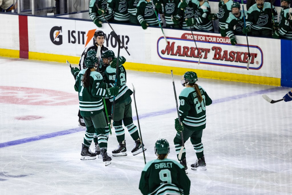 Fleet players celebrate Kluge's first PWHL goal with smiles and a group hug. They are wearing green home uniforms.