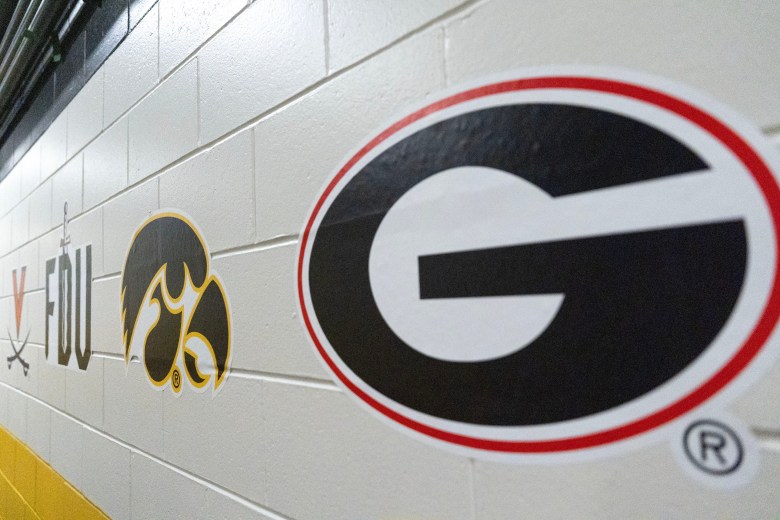 The logos of Georgia, Iowa, Fairleigh Dickinson and Virginia are shown on a white brick wall. Georgia's looks the largest because it's closest to the camera, and Iowa's is the next closest.