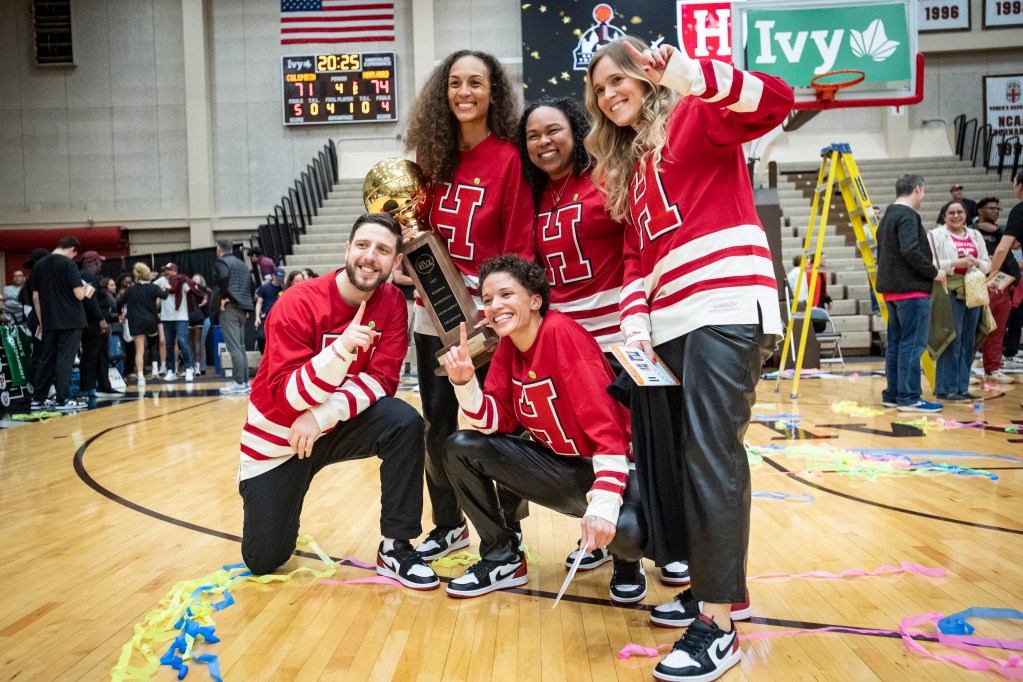 Harvard head coach Carrie Moore, her three assistant coaches and her director of basketball operations smile for a photo with the Ivy League Tournament trophy. Several of them are holding up one finger. They are wearing crimson hockey jerseys with a block “H” on them and black leather pants.