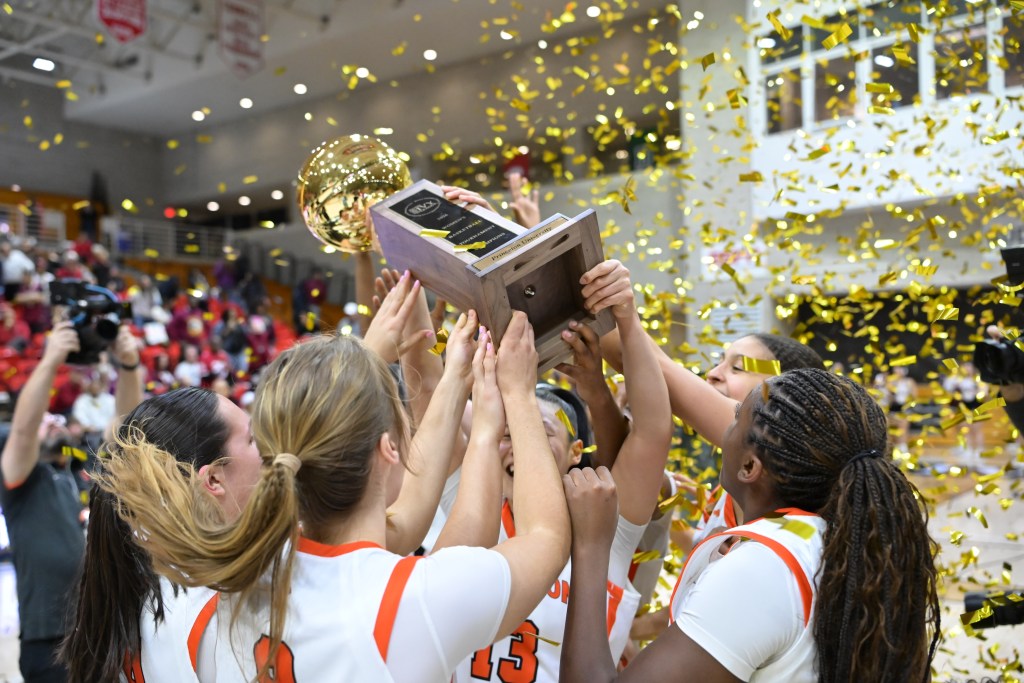 Several Princeton players hold a large trophy with a gold ball at the top over their heads. Gold confetti flies through the air around them.
