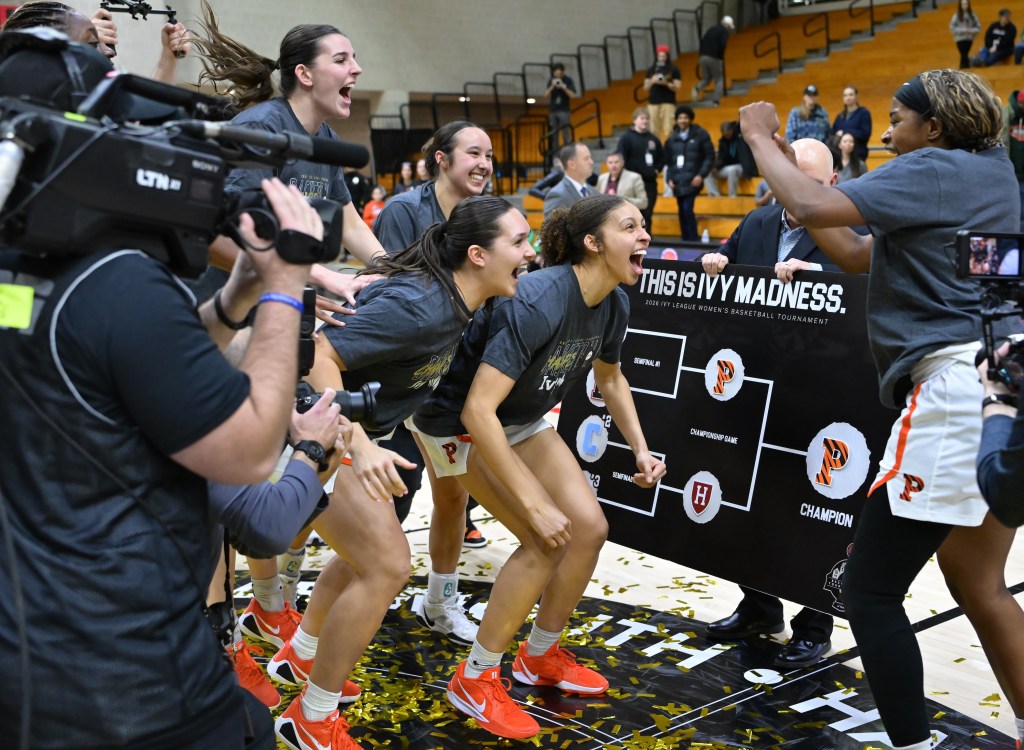 Princeton guard Madison St. Rose shouts in celebration with her teammates after placing the Princeton logo in the "Champion" slot on the Ivy League Tournament bracket. They are all wearing dark gray championship T-shirts, and gold confetti is all over the court.