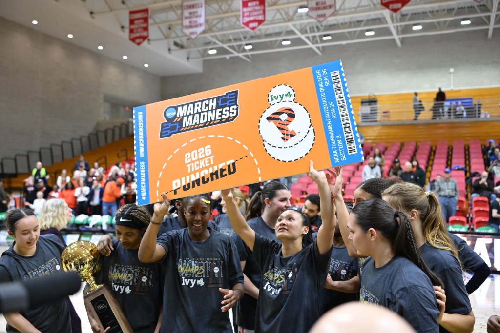 Princeton guard Ashley Chea and guard/forward Olivia Hutcherson hold up an orange piece of cardboard that is decorated like a ticket. It reads, "March Madness, 2026 ticket punched," and has the Ivy League and Princeton logos on it. Chea and Hutcherson's teammates crowd around them, and they're all wearing dark gray championship T-shirts.