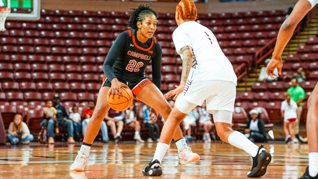 A player in a black uniform dribbles the basketball while a player in a white uniform challenges her. 