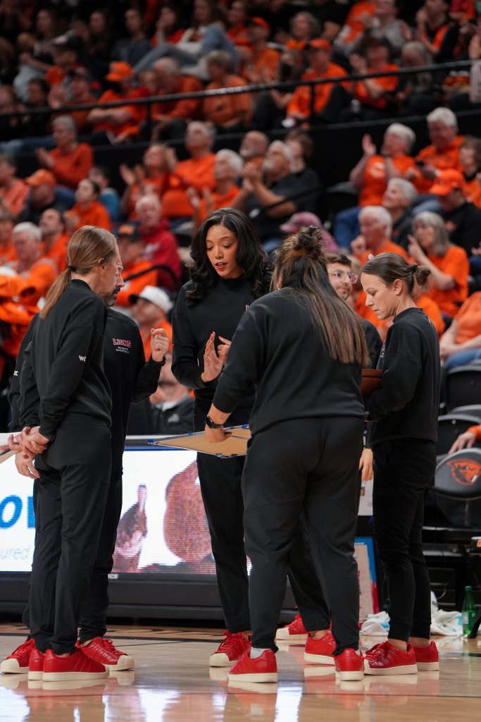 LMU head coach Aarika Hughes talks to her staff during a timeout. She is in the back, facing the camera, with four of her assistant coaches facing her. There is a large but out-of-focus crowd behind them.