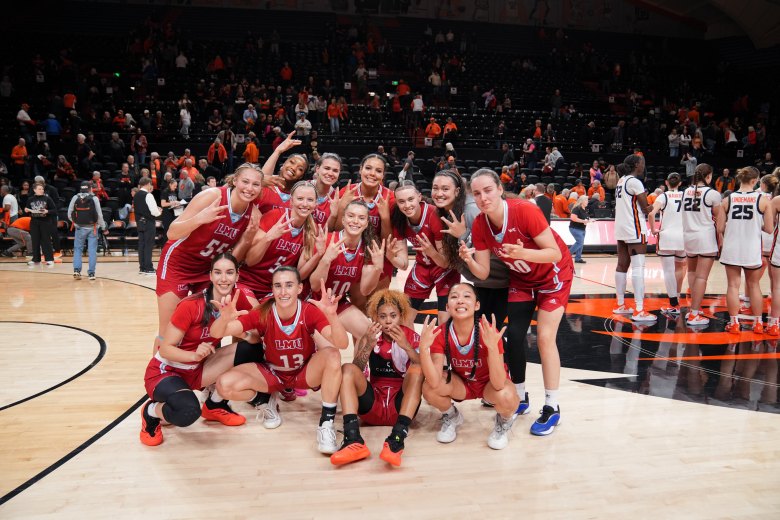 LMU players smile and hold up w's with their hands to celebrate their conference title. They are all facing forward with a crowd behind them.