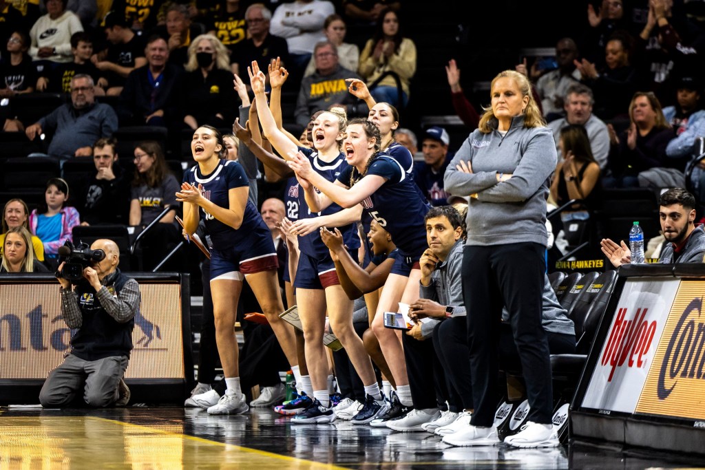 Fairleigh Dickinson head coach Stephanie Gaitley stands in front of her bench with her arms folded across her chest. Her players on the bench shout and put their hands in the air to celebrate a 3-pointer.