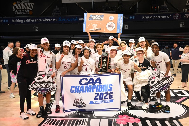 The College of Charleston celebrate its 2026 Coastal Athletic Association championship as players hold trophies and signs. Another in the back holds a large orange placard