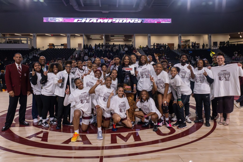 Alabama A&M team members celebrate at midcourt following its first SWAC regular season championship in program history. The coach is holding the trophy in the center of the photo as players and staff smile.