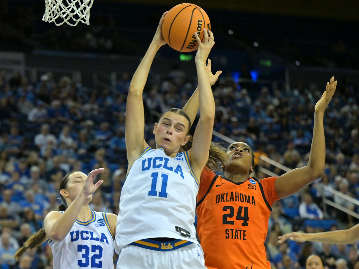 UCLA forward Gabriela Jaquez grimaces as she snatches a rebound with both hands. Oklahoma State’s Praise Egharevba jumps behind her while the a full UCLA crowd is blurred in the background.