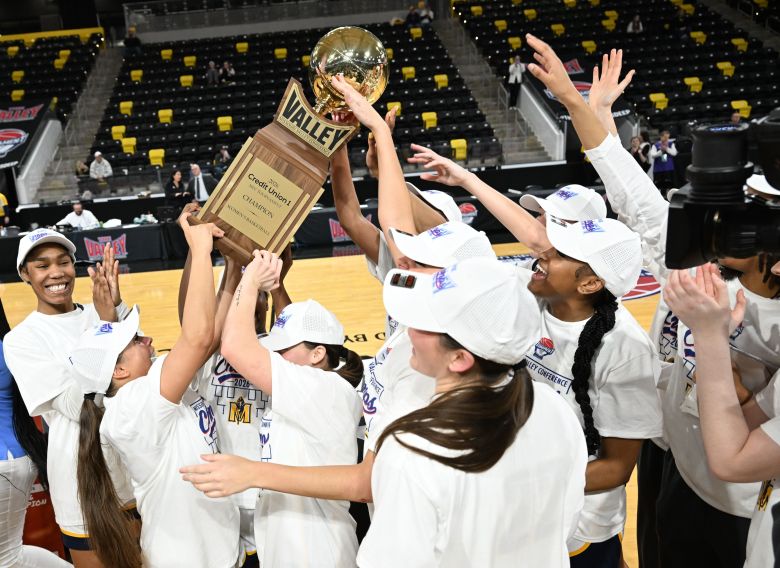 Murray State players lift up the championship trophy after winning their second straight MVC Tournament at XTream Arena in Coralville, Iowa, on March 15, 2026. The players are all wearing white championship t-shirts and hats.
