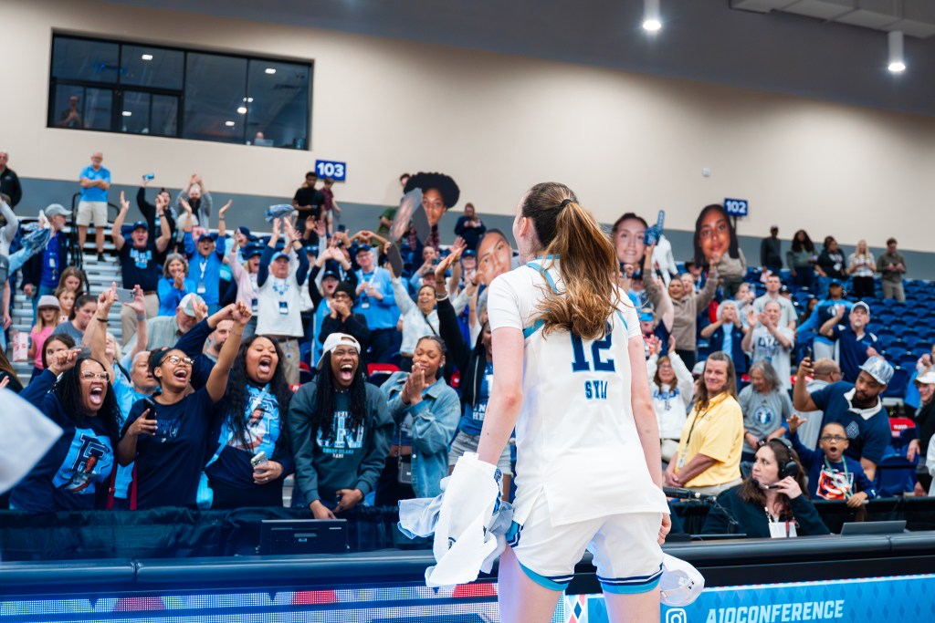 Rhode Island sophomore center Albina Syla is facing the fans with a towel and her championship shirt in her left hand and her championship hat in her right hand. The fans raise fatheads, show heart hands, applaud and cheer.