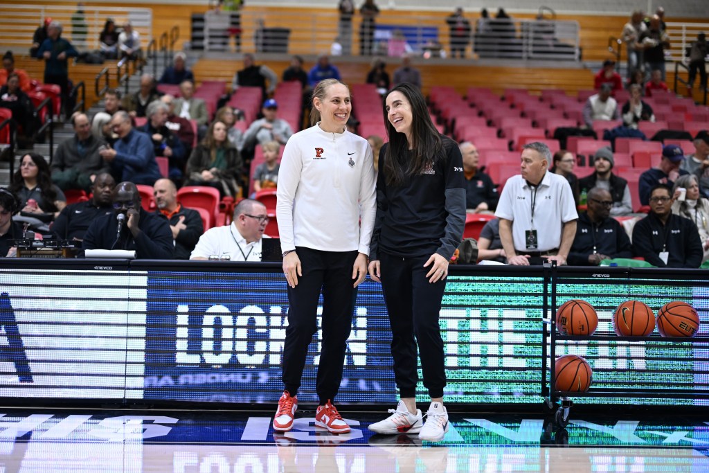 Princeton head coach Carla Berube and Brown head coach Monique LeBlanc stand next to each other on the sideline and talk before a game. Some fans are visible in the stands behind them, and a ball rack with four basketballs on it is next to LeBlanc.
