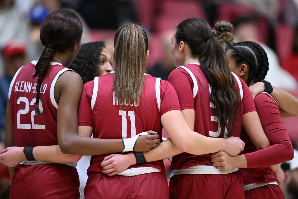 Five Harvard players huddle around head coach Carrie Moore. They are wearing crimson jerseys and have their arms wrapped around one another near each other's waists.