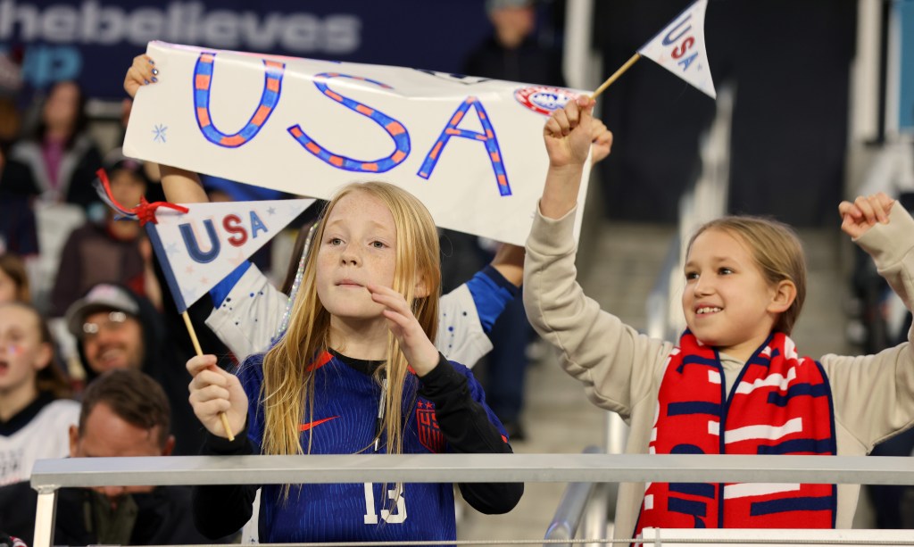 Two young fans hold up a white poster saying "USA" while waving two small white flags. 