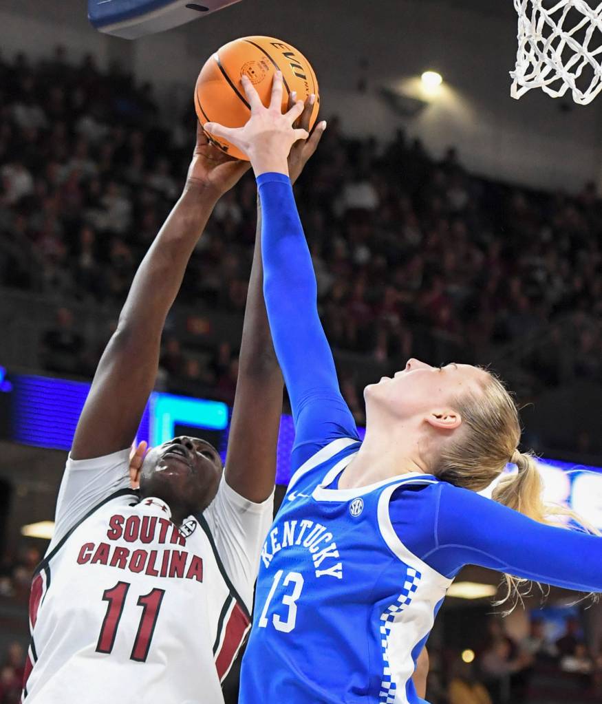 Two players battle for a basketball on a basketball court. One wears. a blue Kentucky jersey and the other wears a white South Carolina jersey.