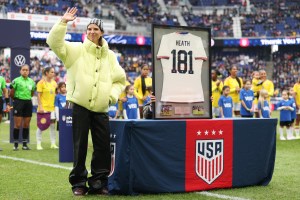 Tobin Heath stands by a table with a US soccer cloth over it and her jersey displayed on top of it with her last name 'Heath' and 181 below it. She waves her hand to greet fans with her other hand in her pocket.