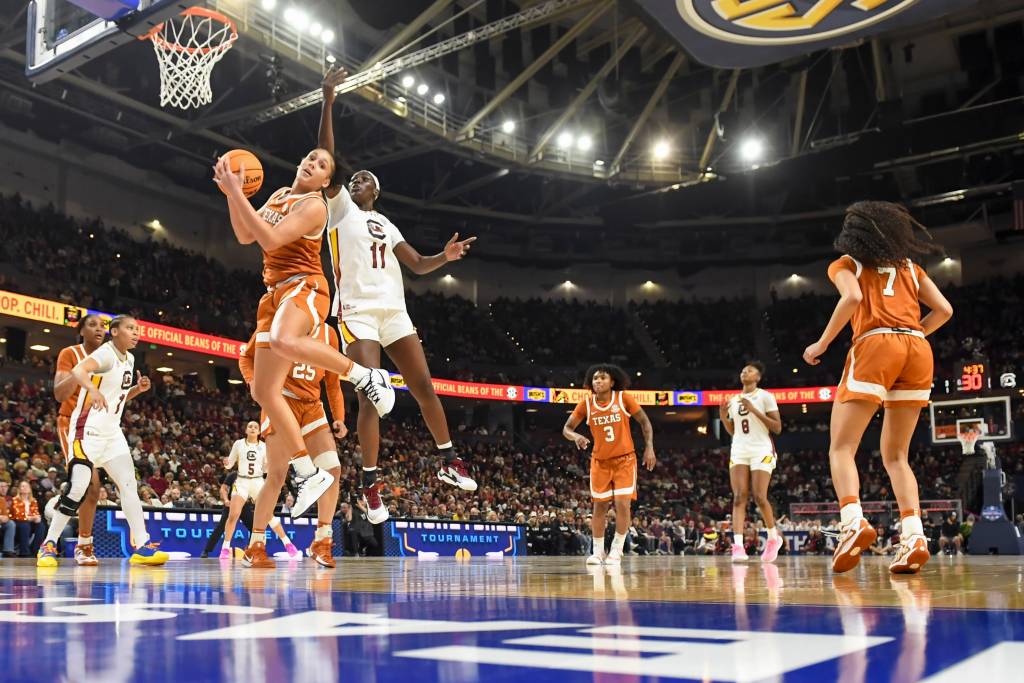 Players battle it out on the court during a basketball game. Five players are in orange jerseys and four are in white jerseys. 