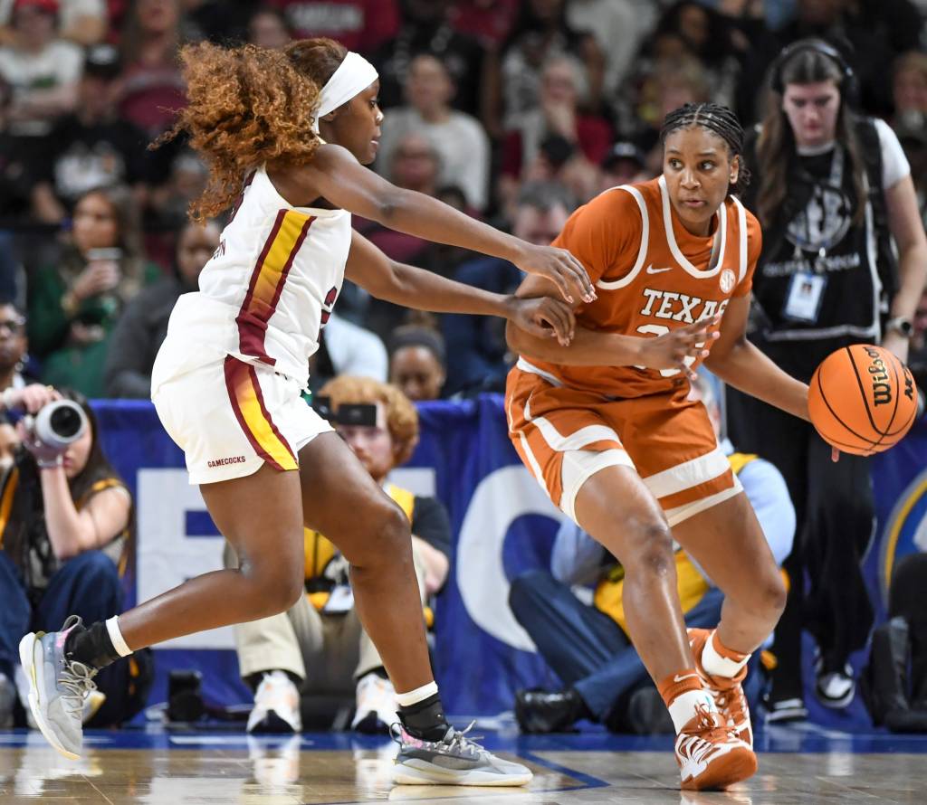 Two players battle for a basketball. One wears a white jersey and the other wears an orange jersey. 