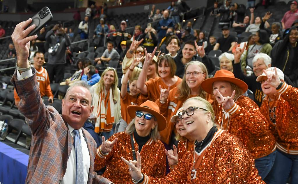 A coach takes a selfie with Texas fans in the crowd, who wear orange glittery jackets. 