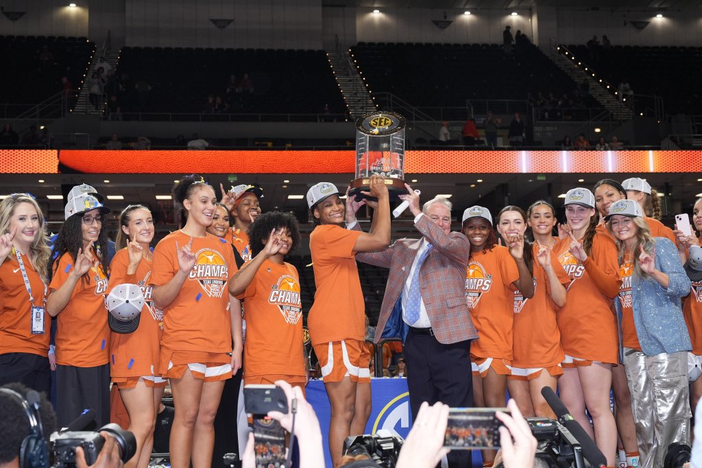 A team poses for a photo after winning a championship. They wear orange and hoist a trophy. 