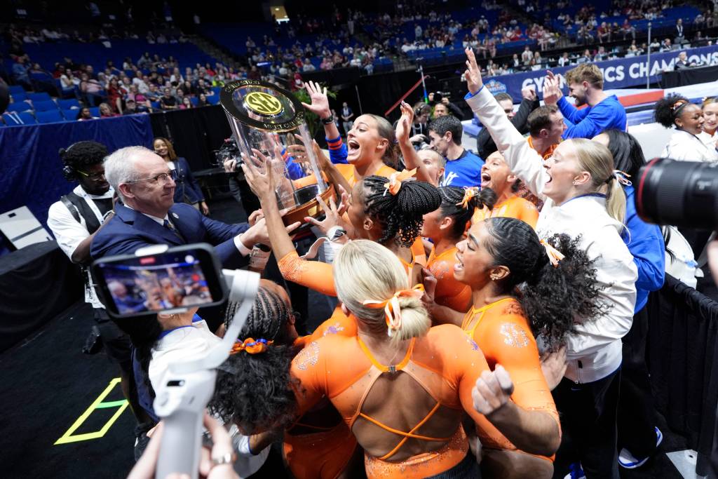 A group of Florida gymnasts in orange leotards celebrate while a man hands them a trophy.