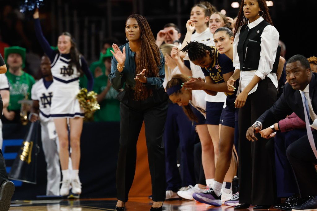 Notre Dame head coach Niele Ivey stands on the sidelines and claps her hands as she watches the action on the court. Her reserves are standing behind her and watching, and some look a little nervous.