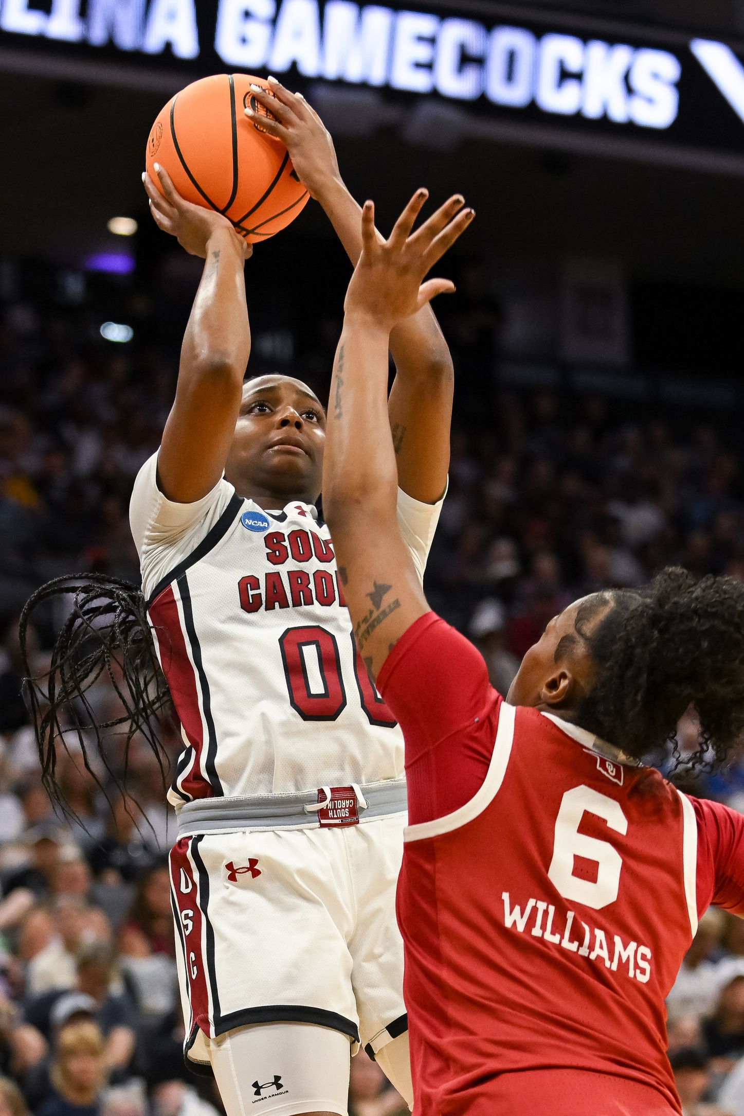 Ta'Niya Latson shoots the ball over an Oklahoma defender. The ball is still in her hands with the Oklahoma defender challenging her shot. Their is an out of focus crowd behind them.