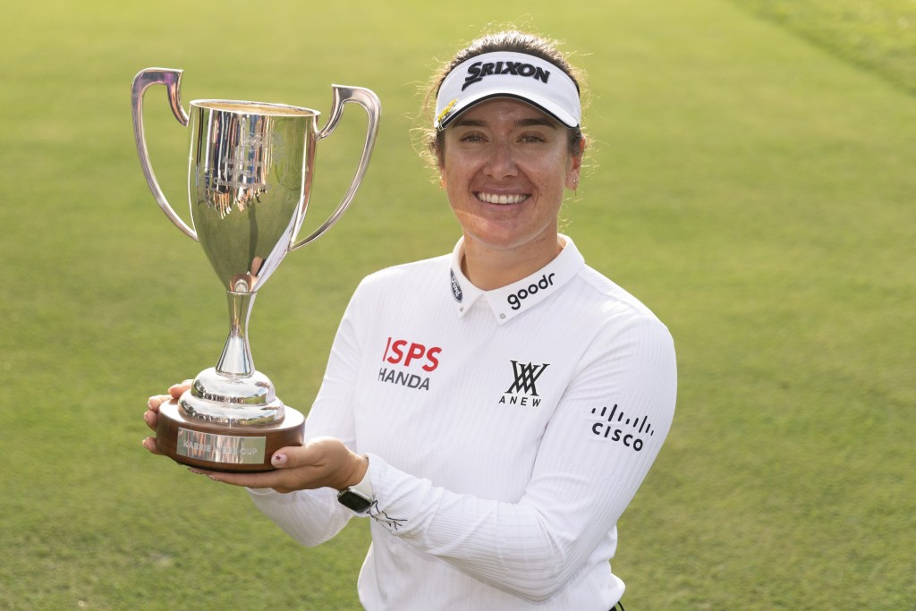 Hannah Green, winner of the 2026 Australian WPGA Championship, holds up the Karrie Webb Cup to her right and smiles. Her hands support the bottom of the trophy, and the background is the green of the golf course.