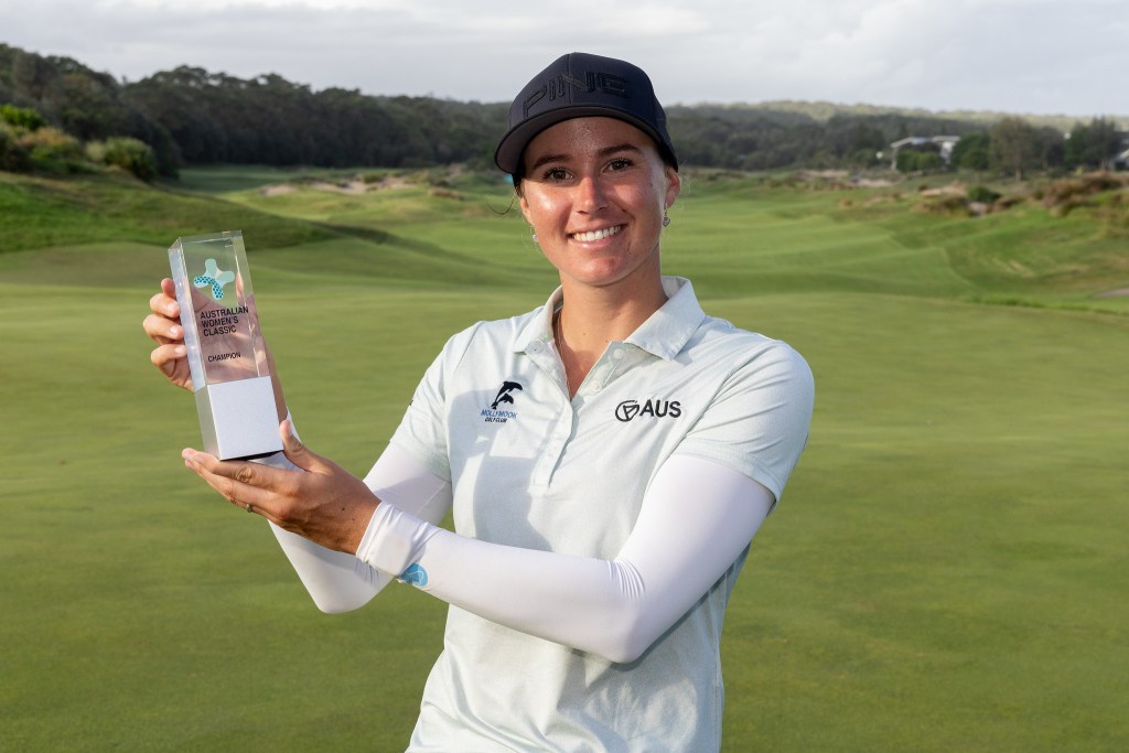 Kelsey Bennett holds up the Australian Women's Classic trophy to her right. She smiles, her right hand is around the back of the trophy and her left hand supports the bottom. The green fairway of Magenta Shores Golf & Country Club stretches behind her.